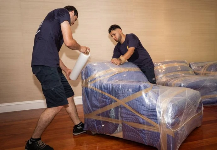 Two movers shrink-wrapping furniture in a living room